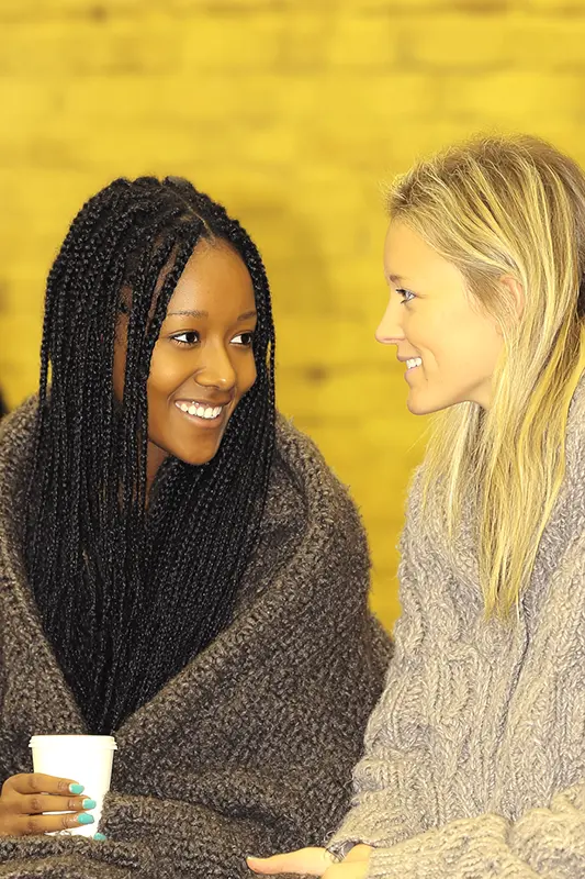 Two people in sweaters warmly converse, one holding a cup, against a yellow brick background, exuding a cozy, friendly atmosphere.