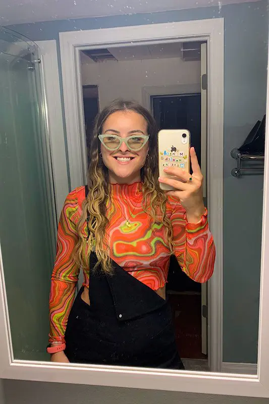 A person with wavy hair and glasses takes a mirror selfie in a vibrant, swirling pattern top, using a smartphone with a colorful case.