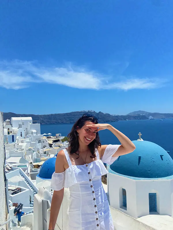 A person in a white dress poses by a blue-domed building in Santorini, Greece, with a bright blue sky and sea backdrop.