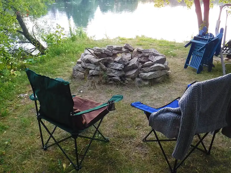 camping Two camping chairs face a stone fire pit by a tranquil lake, surrounded by greenery and dappled sunlight, creating a serene outdoor setting.