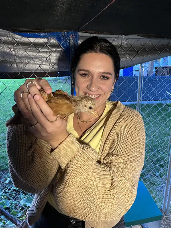A person smiles while holding a small brown bird inside a fenced area, with green grass visible in the background.