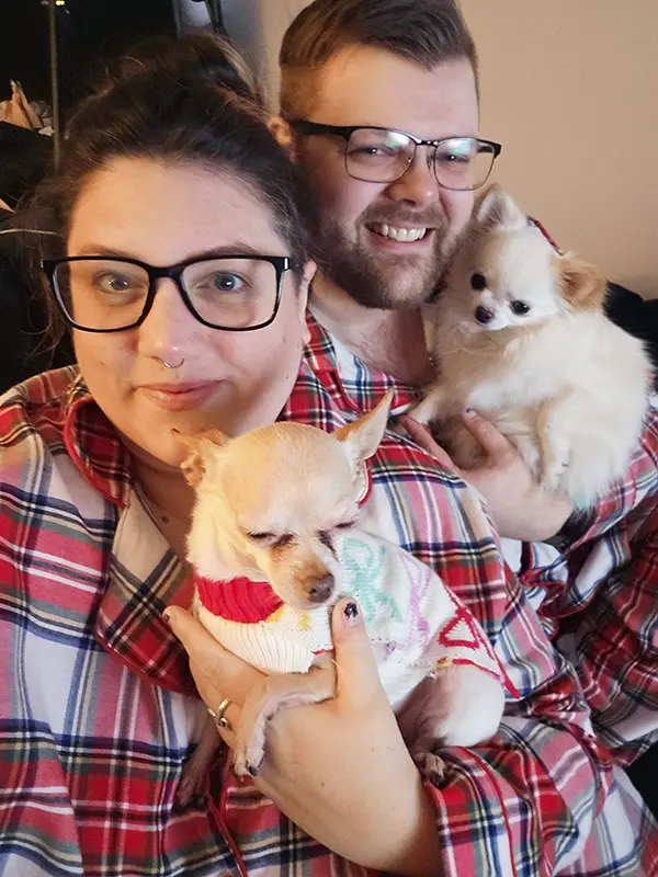 Two people in matching plaid shirts hold small dogs, smiling indoors. Both dogs wear sweaters; one person has glasses.