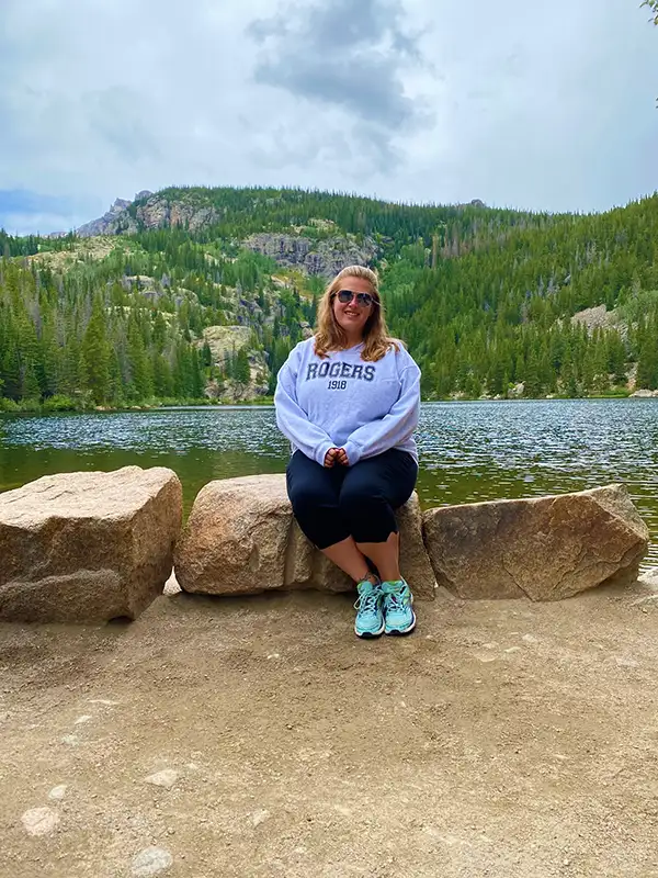 colorado-mountains A person sitting on rocks near Bear Lake, surrounded by lush trees and mountains under a cloudy sky in a serene landscape.