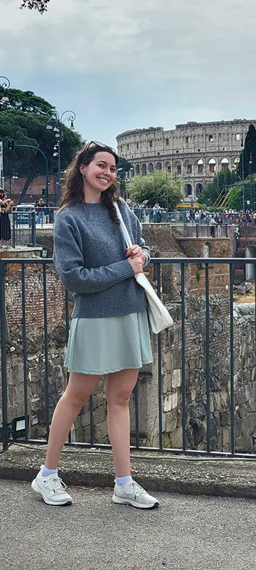 A person stands smiling near a railing, holding an umbrella, with the Colosseum in Rome visible in the background.
