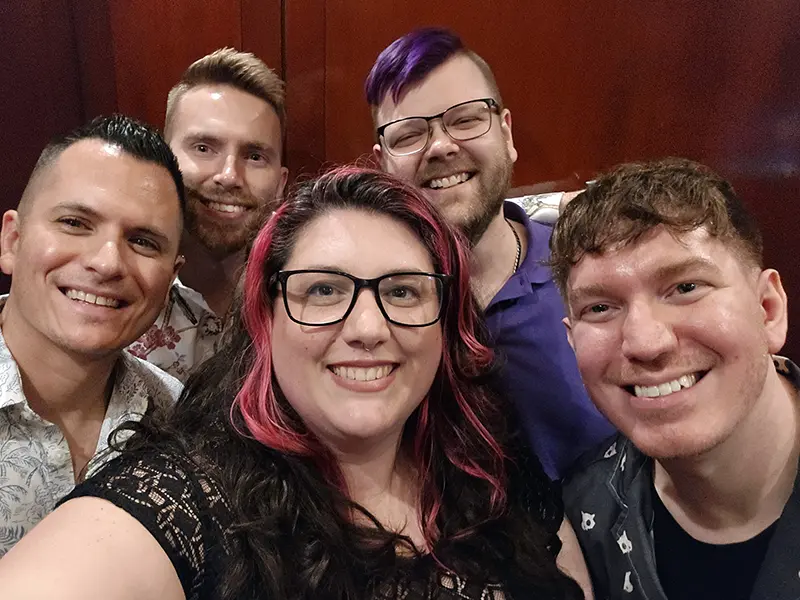 A group selfie showing five smiling individuals in a room with wooden walls. The atmosphere appears friendly and casual. No landmarks visible.