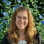 A person with glasses and long hair smiles in front of vibrant blue flowers and green foliage in natural light.