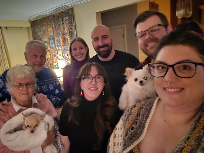 A group of seven people smile together indoors, holding two small dogs. A colorful quilt hangs behind them on the wall.