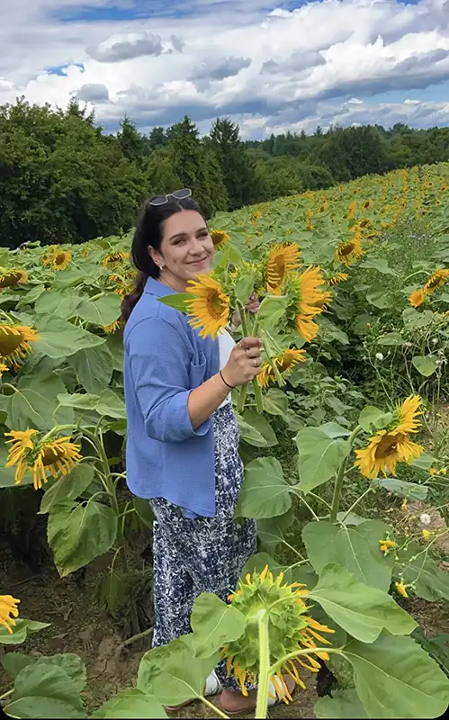 A person in a blue top stands amidst a vibrant sunflower field under a partly cloudy sky, holding sunflowers and smiling.