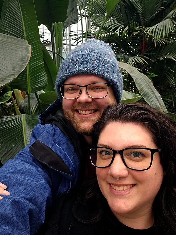 Two people wearing glasses and warm clothing smiling in a lush green tropical setting, surrounded by large leaves and plants.