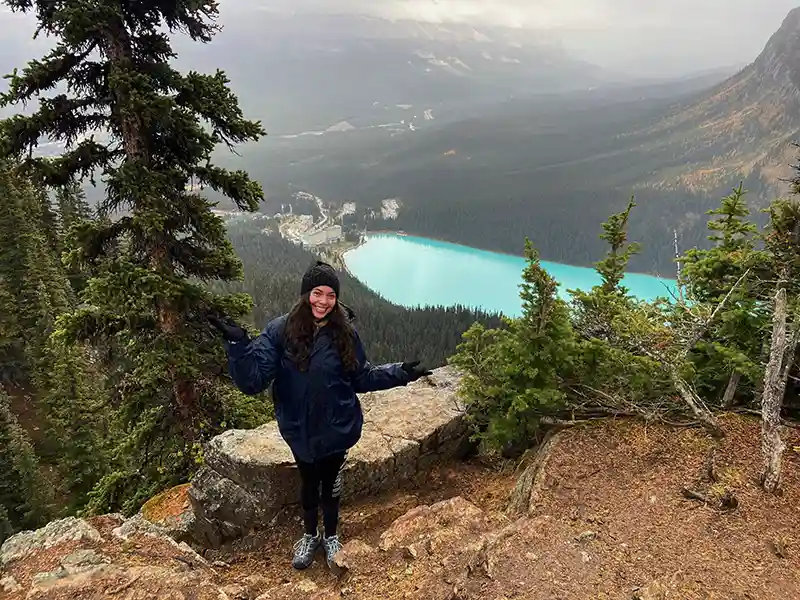 A person smiles on a rocky overlook above turquoise Lake Louise, surrounded by lush forest, with mountains in the background.