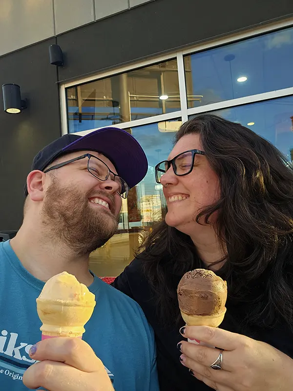 Two smiling people holding ice cream cones, sitting outside a modern building with large windows and reflections. They seem to be enjoying a moment together.