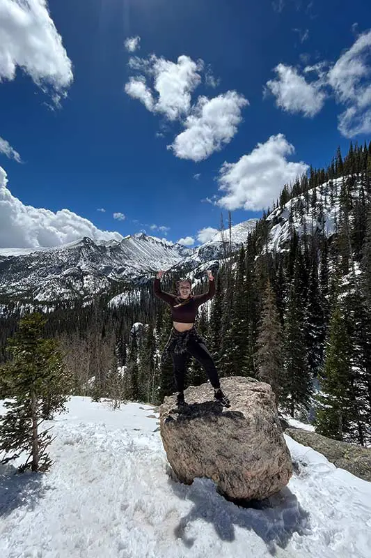 A person poses triumphantly on a rock in a snowy mountain landscape, with clear blue skies and scattered clouds above.