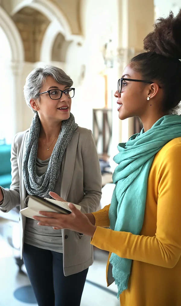 Two people having a conversation inside a building with arched architecture. One is holding a notebook, both wearing glasses and scarves.