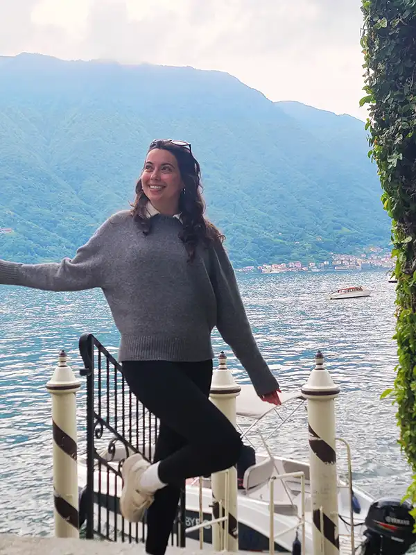 A person smiles by a lakeside railing, surrounded by mountains and a calm lake, with a boat docked nearby.