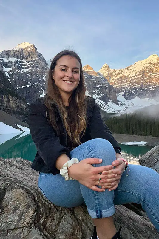 A person sits on a rock by Lake Moraine, surrounded by snow-capped mountains and turquoise waters, smiling at the camera.