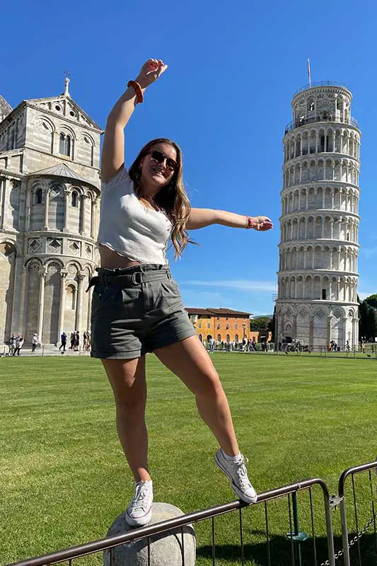 A person playfully poses near the Leaning Tower of Pisa and a cathedral under a clear blue sky, enjoying sunny weather and green surroundings.