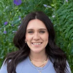 A person with long, dark hair smiles in front of a lush, green background with purple flowers, wearing a light blue top and necklaces.