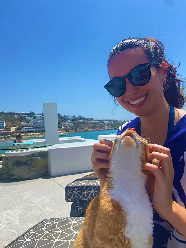 A person wearing sunglasses smiles while petting a cat outdoors, with a coastal landscape and buildings visible in the background.