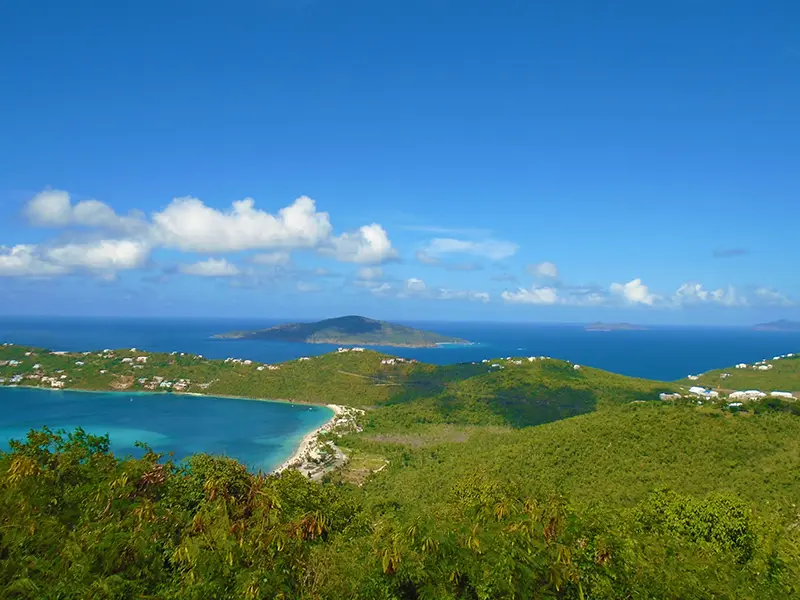 magens-bay Lush green hills overlook a turquoise bay, scattered with small white buildings, under a bright blue sky with distant islands on the horizon.