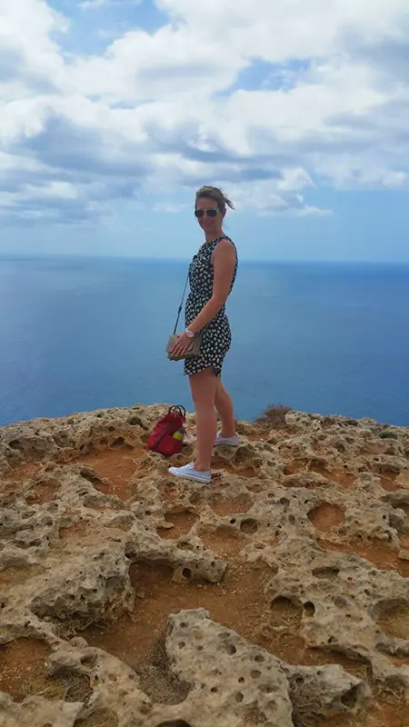 A person stands on rocky terrain overlooking the ocean. The sky is cloudy, and a red backpack is placed on the ground.
