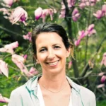 A person smiling in front of blooming pink flowers, set in a lush green outdoor setting, exuding a sense of tranquility.