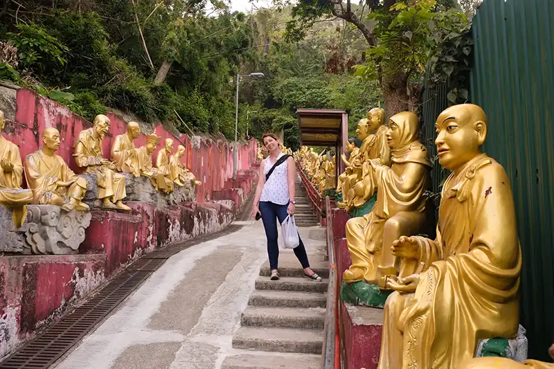 A person stands on stairs surrounded by numerous golden statues of Buddhas at the Ten Thousand Buddhas Monastery in lush greenery.