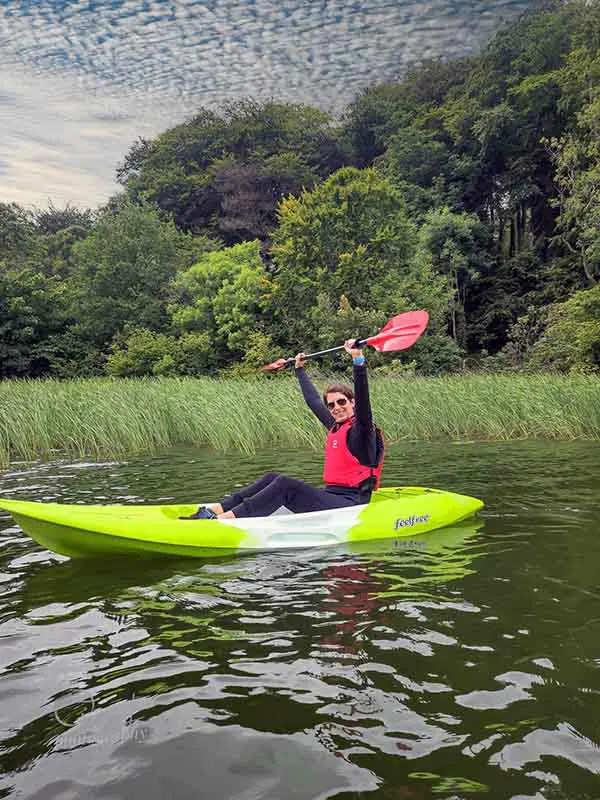 A person in a red life vest holds a paddle joyfully while kayaking on a calm lake surrounded by lush green trees.