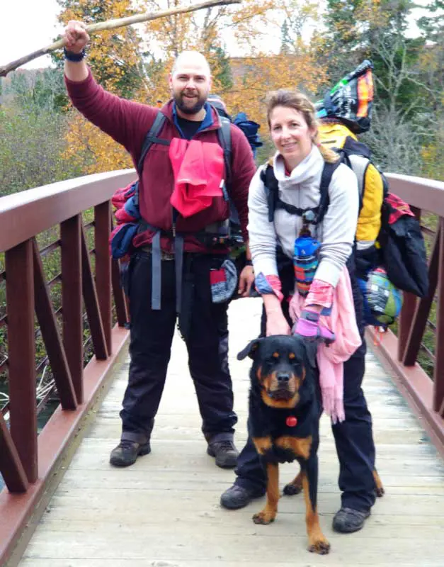 Two people with backpacks and a dog stand on a wooden bridge surrounded by autumn trees, one person holding a stick triumphantly.