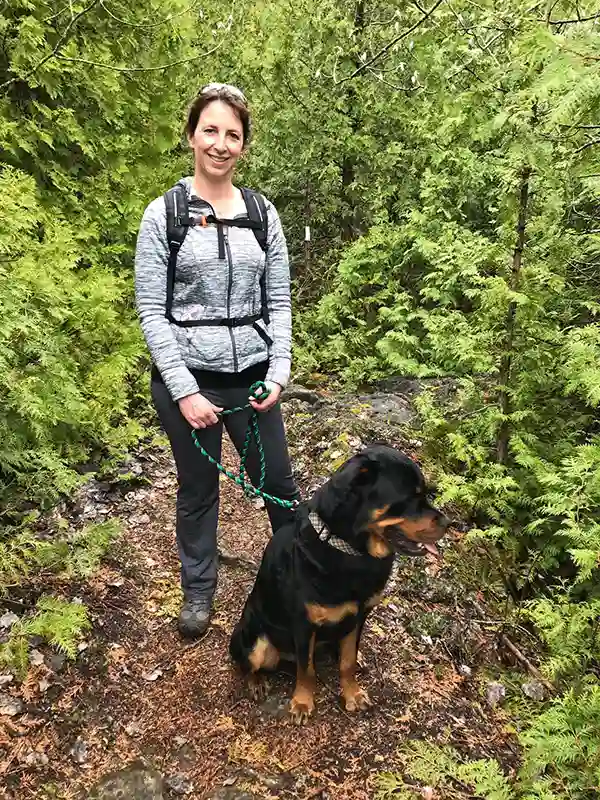 A person stands on a forest trail holding a leashed dog, surrounded by lush green trees and foliage.