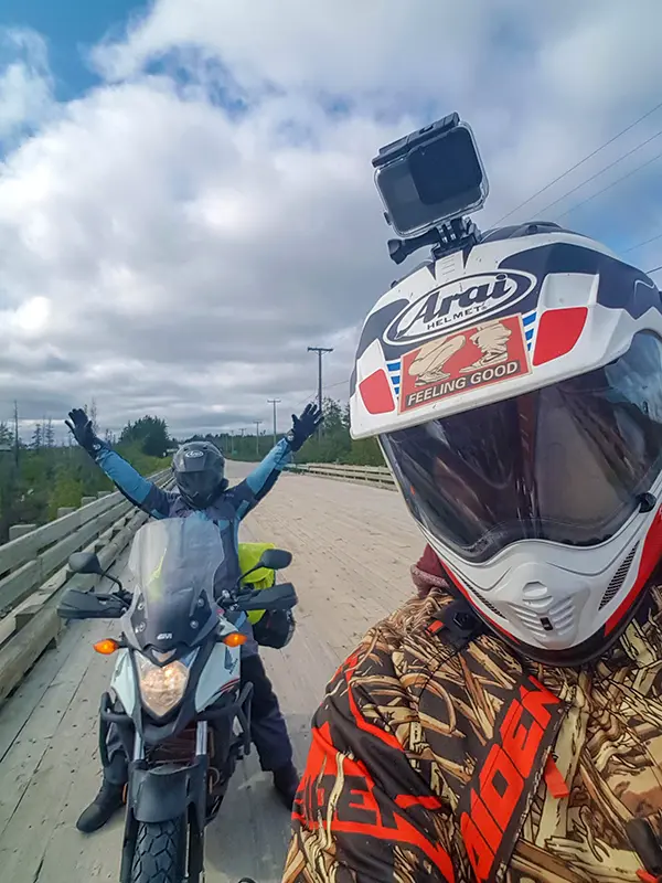 Two people on a wooden bridge with motorcycles. One raises arms, while the other takes a selfie. Overcast sky and trees in background.