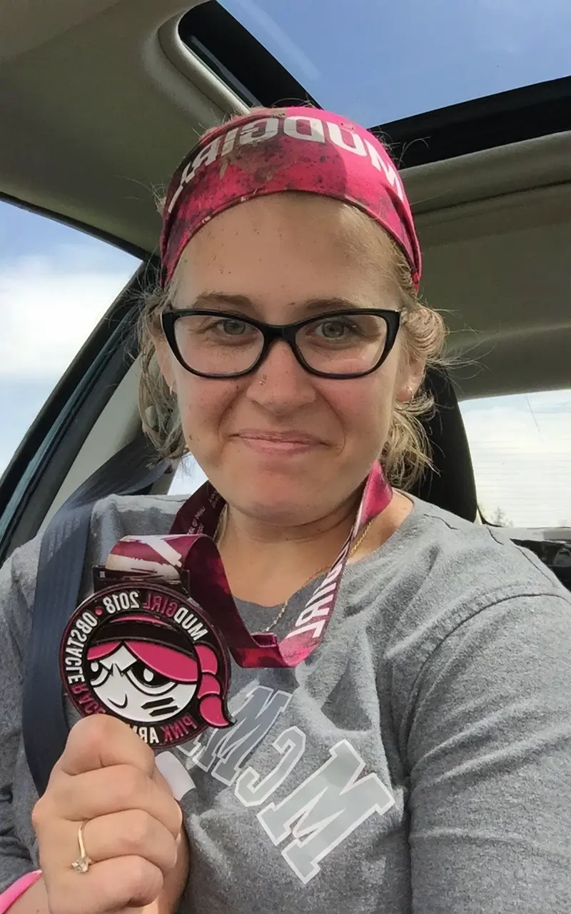 mudgirl-race A person wearing glasses and a pink headband holds a medal inside a car, with a visible sunny sky through the window.