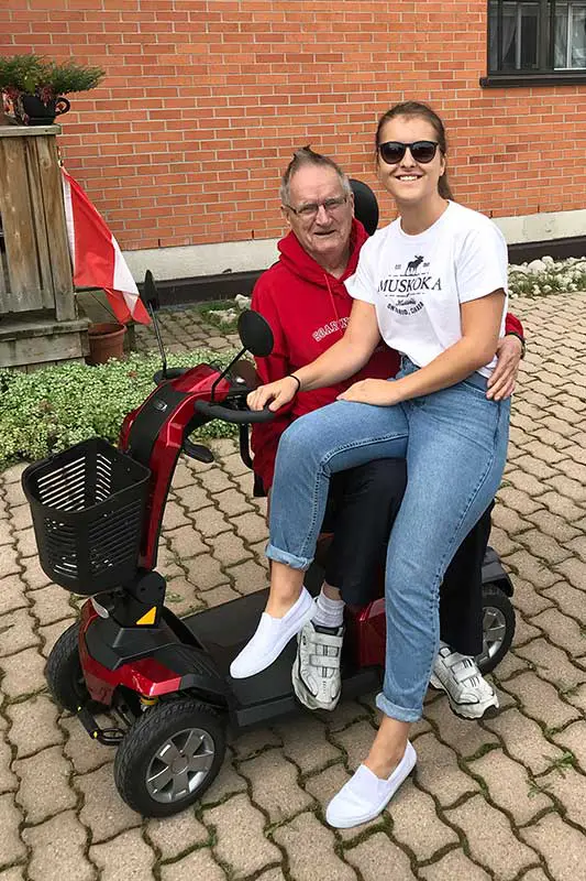 Two people smiling on a red mobility scooter, with a Canadian flag nearby, in front of a brick building.