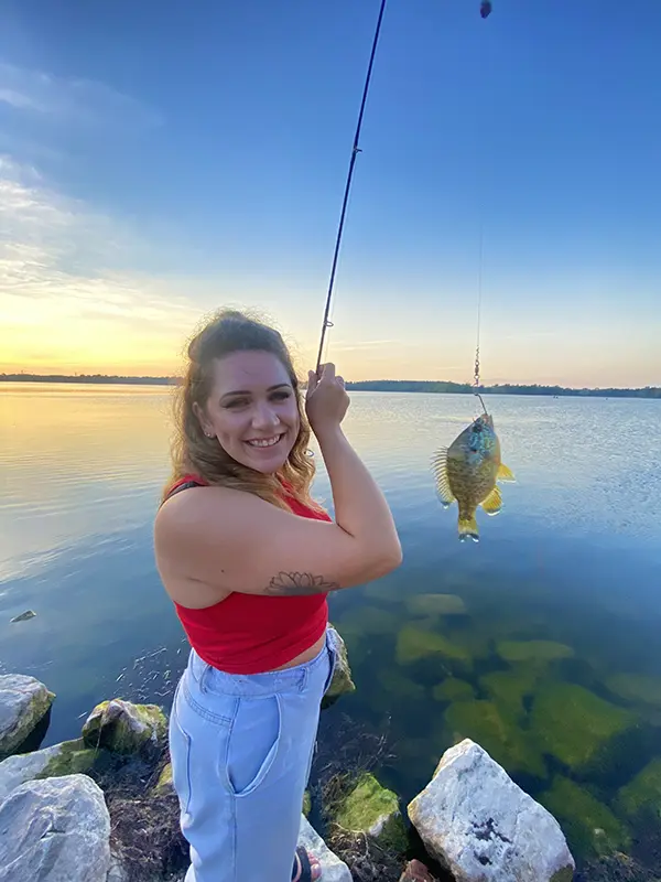A smiling woman in a red top holds a fishing rod with a fish near a calm lake at sunset.