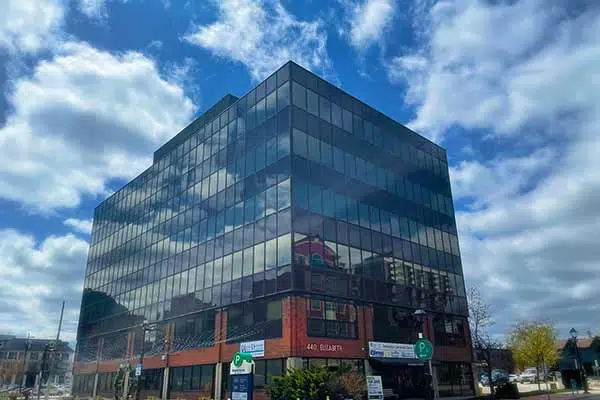 Modern glass building with reflective windows, set against a partly cloudy sky. Street signs and parked cars are visible in the foreground.
