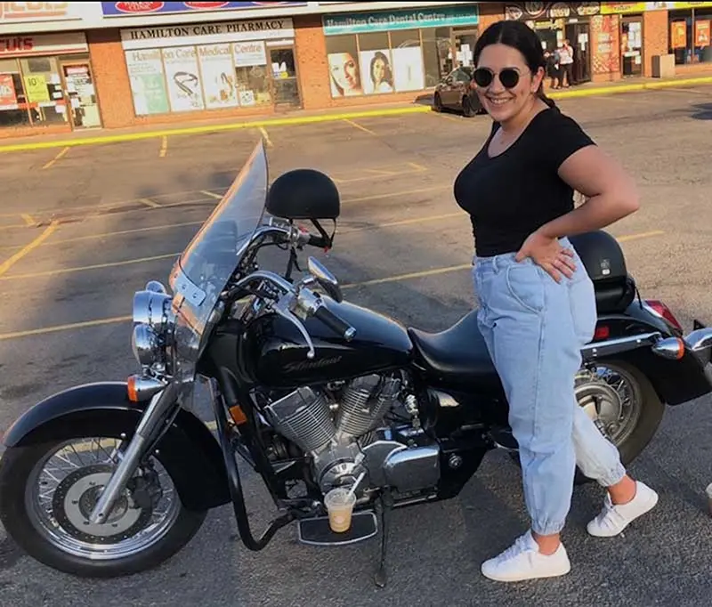 A person in sunglasses stands beside a parked motorcycle in a shopping plaza parking lot, with a drink placed on the bike's footrest.