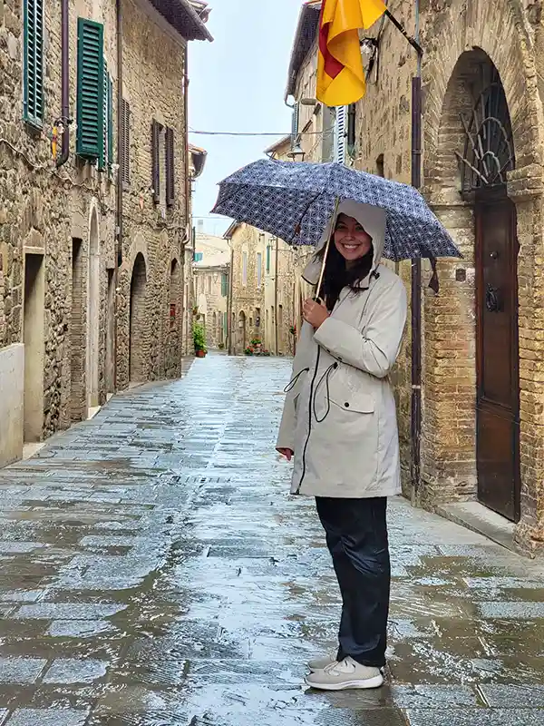 A person holding an umbrella smiles on a rainy cobblestone street within a historic Italian village, surrounded by stone buildings and colorful shutters.