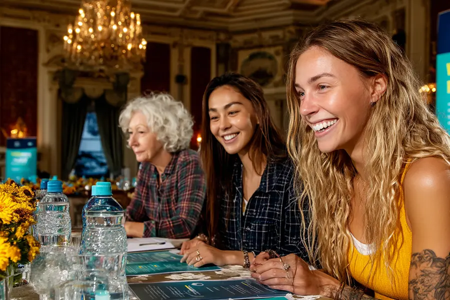 Three people smiling and talking at a table with water bottles and papers in an elegant room with chandeliers and ornate decorations.