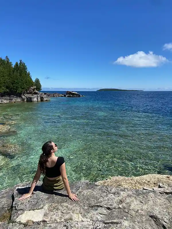 A person sits on rocky shore, enjoying sunny day by the clear blue water under a bright sky with distant island view.