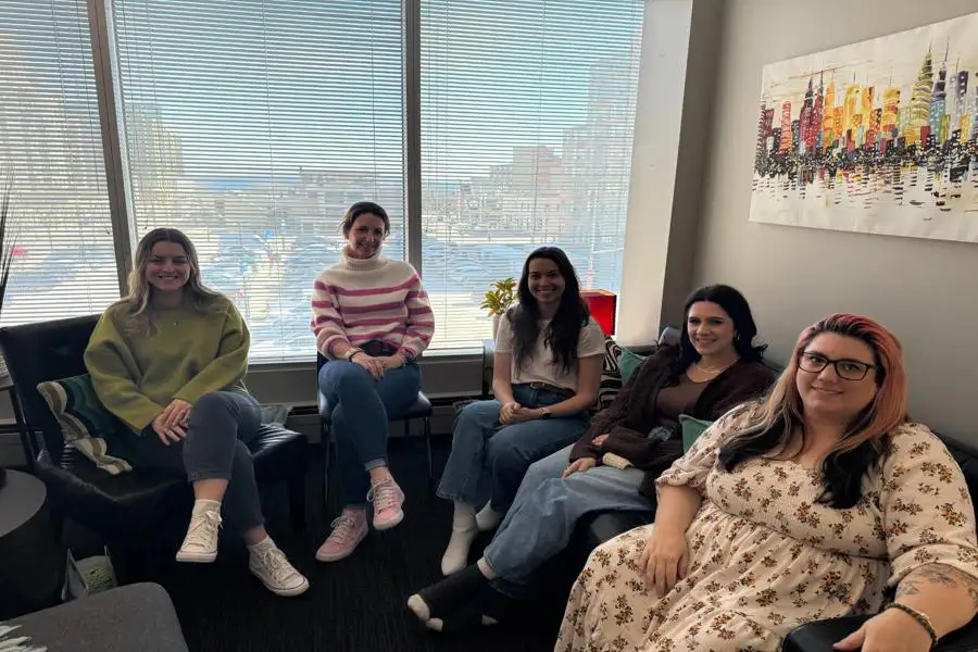 Five people sitting in a cozy room with a cityscape painting. Large window reveals a bright city view outside.