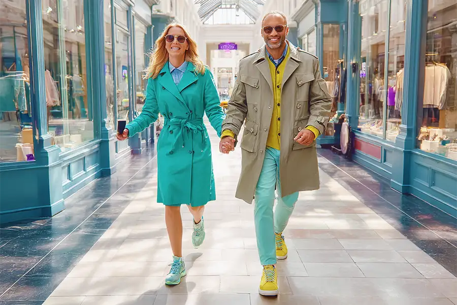 Two people in colorful coats walk happily through a sunlit shopping arcade, surrounded by stores featuring bright, elegant displays.