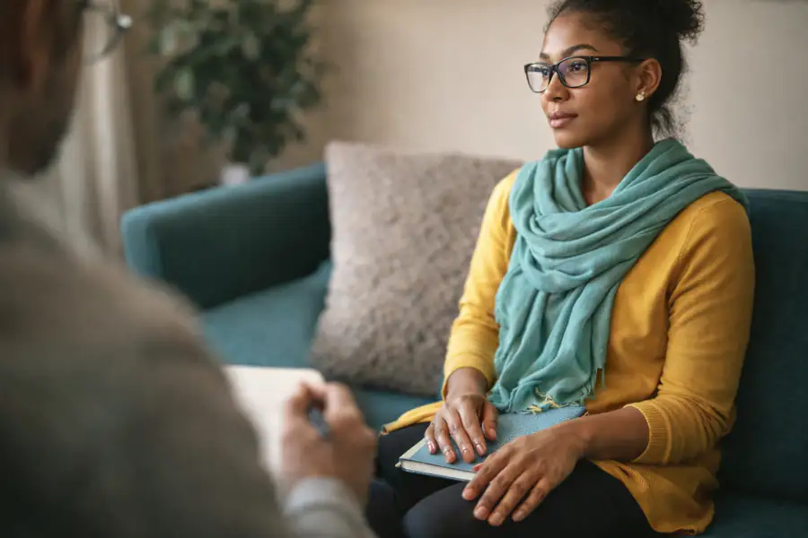 A client sits calmly on a teal couch during a therapy session, holding a notebook, while a therapist listens in the foreground.