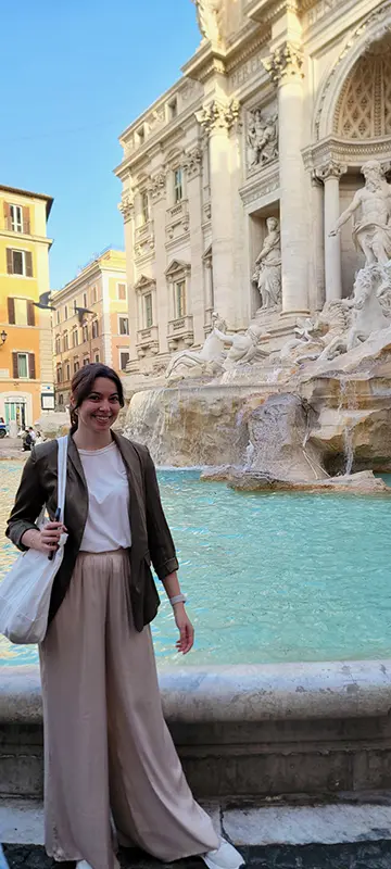 A person stands smiling beside the Trevi Fountain in Rome, with sculptures and cascading water visible against an old building backdrop.
