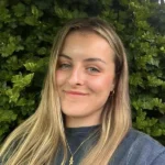 A person with long hair stands in front of dense green foliage, wearing a necklace and earrings, smiling at the camera.
