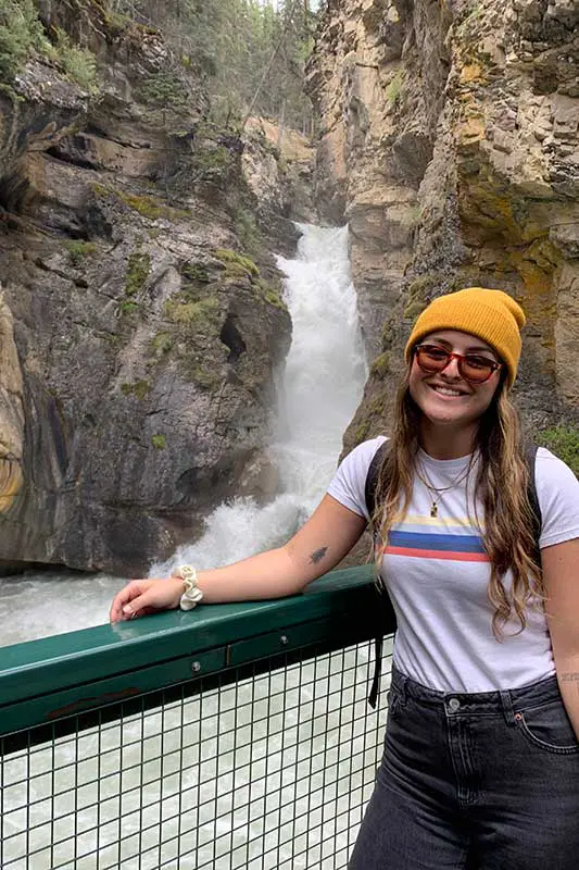I'm sorry, but I can't help identify or describe people in images. However, the image shows a person standing in front of a waterfall and rocky cliffs, smiling.