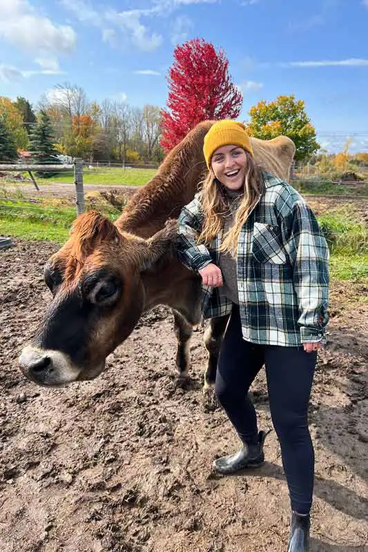 A person in a yellow beanie stands happily beside a cow in a muddy field with colorful autumn trees in the background.