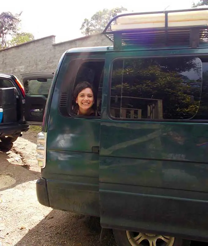A smiling person looks out a green van's window in a sunny, outdoor setting with parked vehicles and a stone wall nearby.