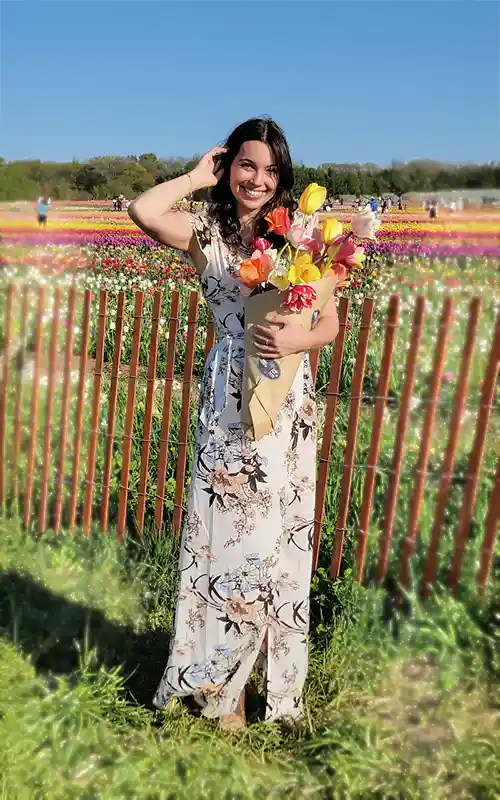 A person in a floral dress holds a bouquet in a vibrant tulip field, with a wooden fence and trees in the background.