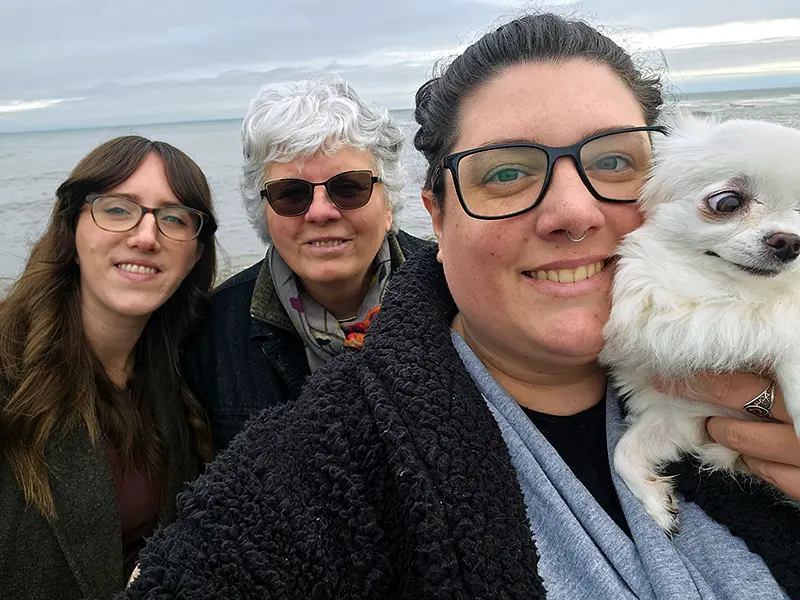 Three people pose with a small dog by the sea, smiling on a cloudy day. No landmarks or historical buildings are visible.