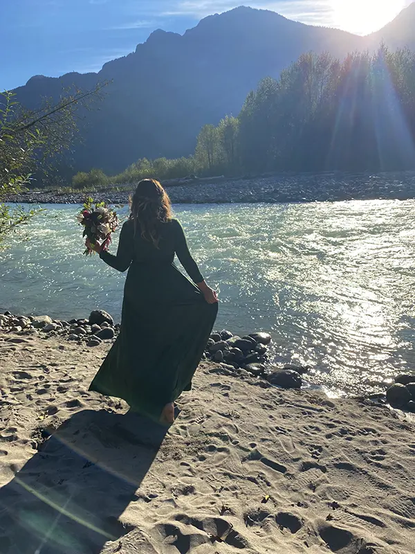 A person in a green dress holds flowers, standing by a river under mountains, with sunlight streaming through the trees.